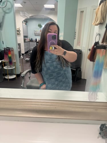 Reviewer in a hair salon takes a mirror selfie wearing the blue, patterned The Whitmore apron.