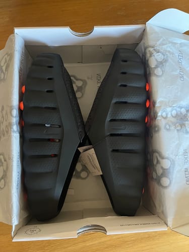 Un client montre les claquettes noires Nike Mind 001 dans leur boîte, avec la semelle extérieure texturée visible.