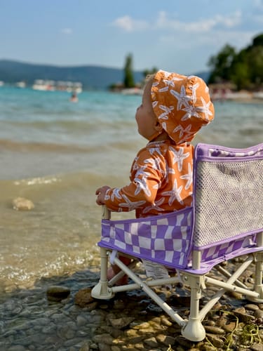 Customer's baby sitting in a purple checkered Ava baby chair on a rocky beach looking at the water.