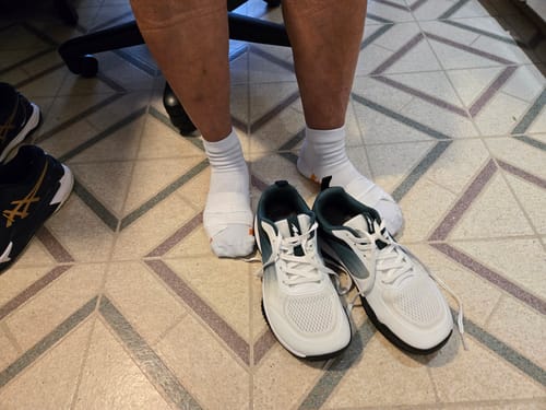 Customer sitting with their feet in socks next to a pair of white CUURT Muuv court shoes on a tiled floor.