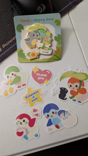 Reviewer's Helping Hand Pin in its packaging, displayed on a table with several matching stickers.
