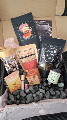 Ein Rezensent zeigt die geöffnete Witchy Box Juli mit einem Trinkglas mit Halloween-Motiven und verschiedenen magischen Artikeln.