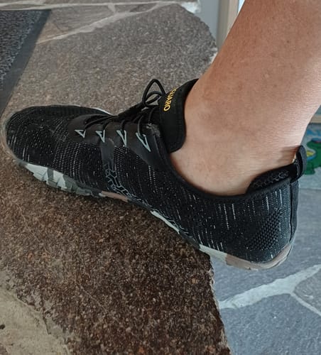 Kunde trägt den schwarzen, gemusterten Smart II Barfußschuh am Fuß auf einer Steinstufe.