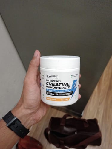 Reviewer's hand holding a white tub of Wellcore Micronised Creatine Monohydrate, with a gym bag in the background.