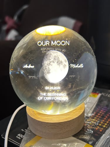Customer's personalized crystal orb showing an engraved moon, names, and a date, illuminated by its wooden base.
