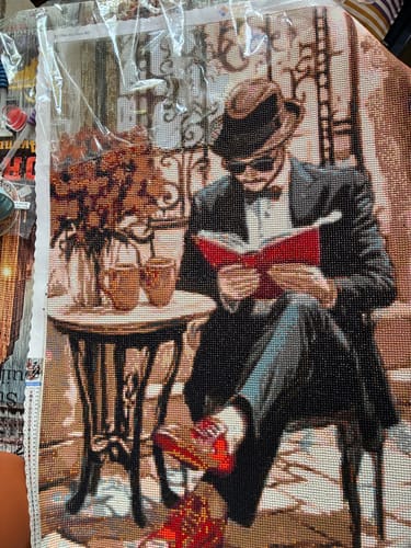 Kunde zeigt sein fertiges Diamond Painting eines Mannes mit Hut, der in einem Café ein Buch liest.