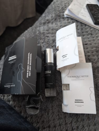 Customer's Smoke & Mirrors Eau de Parfum bottle with its black box and paper samplers on a grey surface.