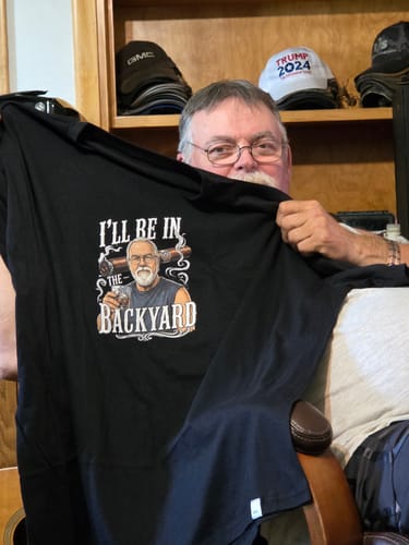 Customer holding up his black 'I'll Be In The Backyard' t-shirt with a personalized cartoon graphic.