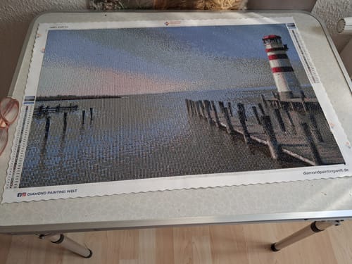 Ein Kunde zeigt sein fertiges Diamond Painting eigenes Bild mit einem Leuchtturm und einem Steg am Wasser.