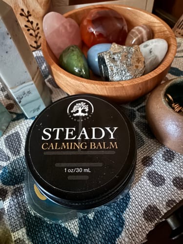 Customer's tin of Steady Calming Balm sits on a patterned cloth next to a wooden bowl of crystals.