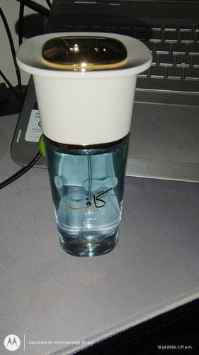 Cliente mostrando el perfume KAAF en su frasco de vidrio con líquido azul y un gran tapón blanco y dorado.