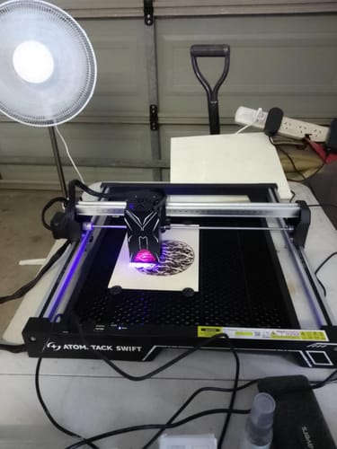 Customer's black AtomStack F2N Honeycomb panel being used with a laser engraver to cut a circular design on a wood square.