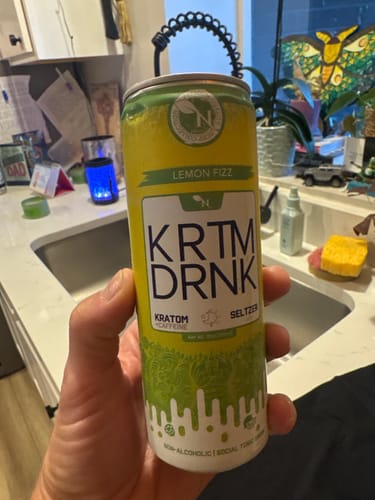 Customer holding a yellow and white can of KRATOM BEVERAGE - LEMON FIZZ in a kitchen.