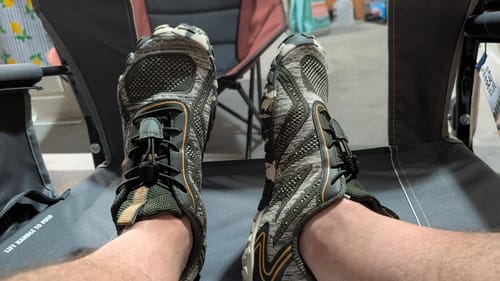 Reviewer's perspective of their feet in camouflage Active II Pro - Barefoot Shoes while sitting in a chair.