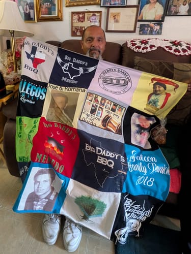 A reviewer sits on a couch, holding up a large Project Repat T-Shirt Quilt made of many custom family shirts.