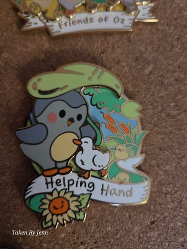 Reviewer's close-up of the colorful Helping Hand Pin on a corkboard, showing its gold details and cartoon bird design.