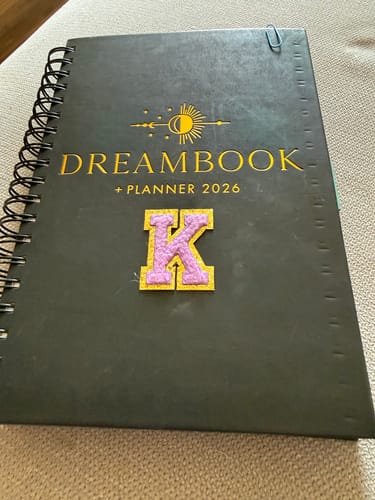 Reviewer's dark grey, spiral-bound Dreambook + Planner 2026, customized with a purple and yellow letter 'K' patch.