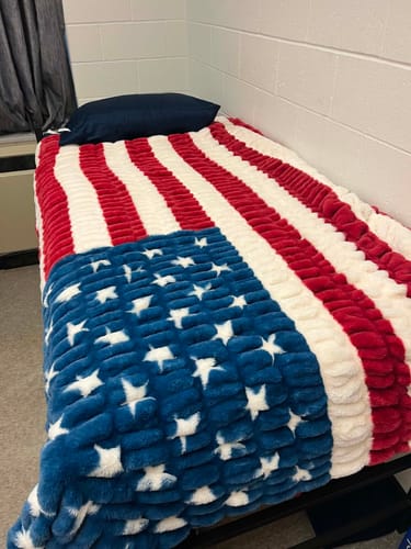 Reviewer's plush Hugs Flag Blanket spread over a single bed in a barracks room.