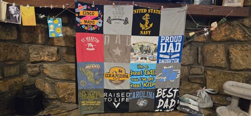 Customer's memorial Project Repat T-Shirt Quilt, featuring shirts with 'Proud Dad' and 'Navy', hanging on a stone wall.
