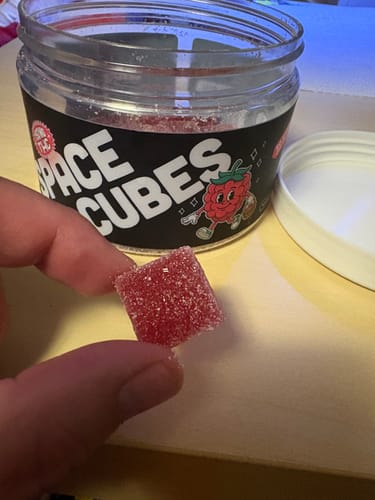 Ein Kunde hält einen roten, gezuckerten THC Space Cube, im Hintergrund ist die geöffnete Dose zu sehen.