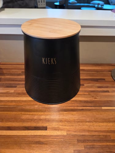 Kundens svarte oppbevaringsboks for kjeks med bambuslokk står på en trebenk.