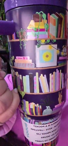 Close-up of a reviewer holding the purple Thicc Buddy 'Shelf Love' tumbler with a colorful bookshelf design.