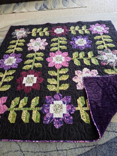 Customer's finished Edelweiss Quilt with colorful batik flowers on a dark background, showing the plush purple backing.