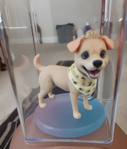 Customer's custom chibi figure keychain of a light-brown dog with a yellow bandana, shown inside a clear display case.