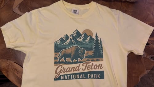 Customer's pale yellow Grand Teton Retro T-Shirt laid flat on a wooden surface, showing the bison and mountain graphic.