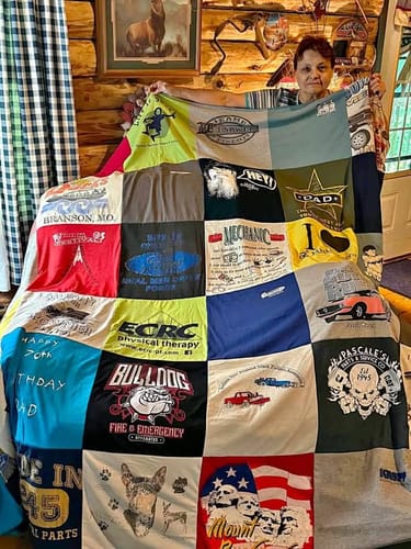 Customer holding up a large, colorful Project Repat T-Shirt Quilt made from many different t-shirts in a living room.