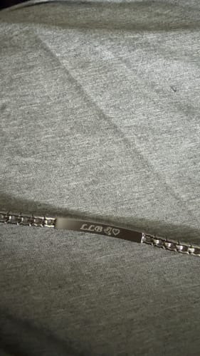 Close-up of a customer's engraved ID Tennis Bracelet on a grey fabric, showing the initials LLB, a dove, and a heart.