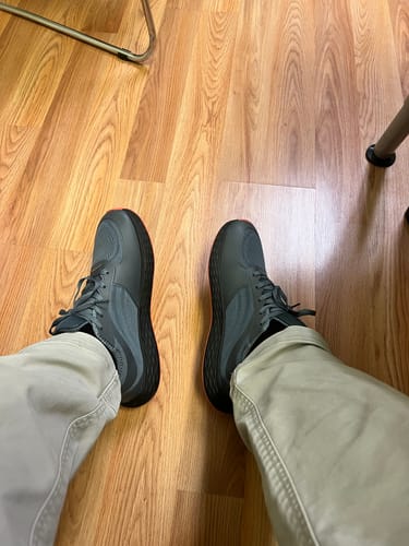 Customer's top-down view of their feet wearing the dark gray CUURT Muuv court shoes on a wood floor.