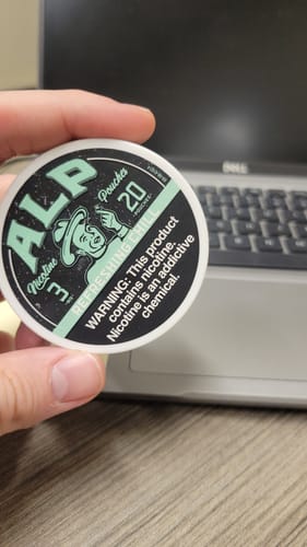 Reviewer's hand holding a circular tin of Refreshing Chill Nicotine Pouches in an office setting.