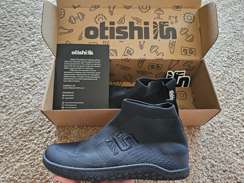 Customer holding a black, knit Otishi 2.0 shoe in their hand, with the open product box in the background.