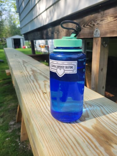 Customer's custom vinyl sticker on a blue water bottle resting on a wooden plank outdoors.