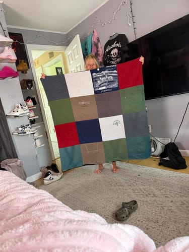 Customer holding up their finished Project Repat T-Shirt Quilt, made from various t-shirt squares, in a bedroom.