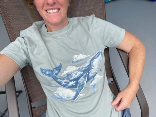 Customer smiling while wearing the light green Whale Above T-Shirt with a whale and clouds graphic.