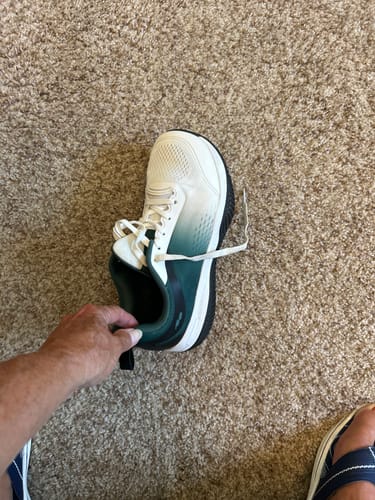 Reviewer's hand holding a white and teal CUURT Muuv court shoe, seen from above on a carpet.