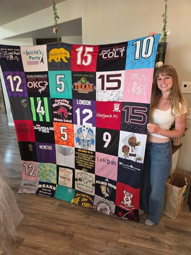 A reviewer smiles while holding up their large Project Repat T-Shirt Quilt made from colorful sports jerseys and t-shirts.