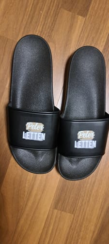 Kunde zeigt ein Paar schwarze, personalisierte Deine Letten Badeschlappen auf einem Holzboden.