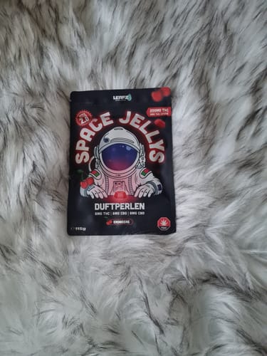 Rezensent zeigt die schwarze Verpackung der THC Spacejellies 2.0, die auf einem flauschigen, grauen Teppich liegt.