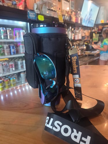 Customer's black Thicc Buddy Crossbody Bag holding a tumbler on a wooden bar, with sunglasses in the front pocket.