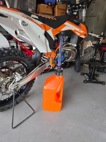 Customer's orange 2.5 Gallon Jug with its spout inserted into the tank of a dirt bike in a garage.