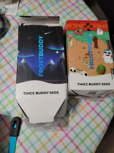 Reviewer's two Thicc Buddy 50oz boxes, one with a Nightfall design and another with a cartoon beach theme, on a table.