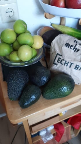 Kundenfoto der grünen Sommer-Zitronen in einer Glasschale neben mehreren Avocados auf einem Holztisch.