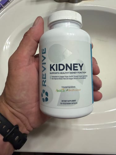 A reviewer's hand holding a white bottle of Revive Kidney supplement over a sink.