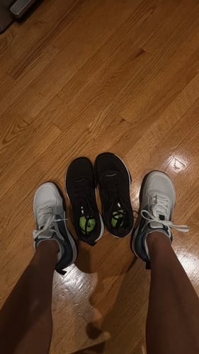 Customer comparing the wide CUURT Muuv shoes in white to a pair of narrower black shoes on a wood floor.