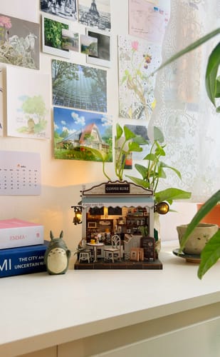 Customer's assembled The Coffee Stop miniature kit, illuminated and displayed on a white desk next to books.