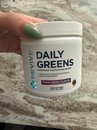 Customer's hand holding a white tub of Revive Daily Greens Powder in the Fresh Berry flavor.