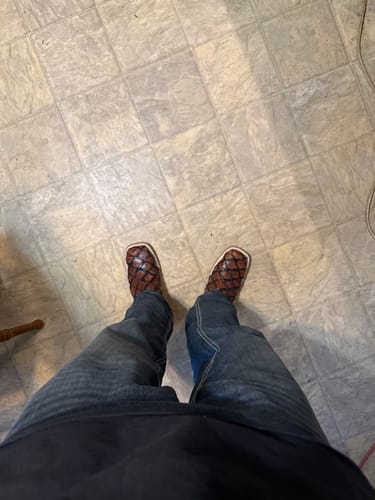 Reviewer looking down at their Cognac Jumbo Pirarucu Leather Boots, worn with jeans on a tiled floor.